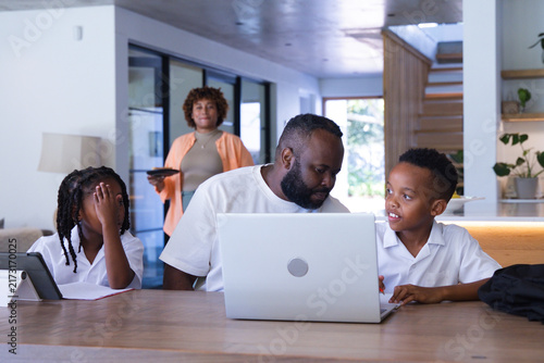 African American family gathering at wooden table in modern home, father working on silver laptop