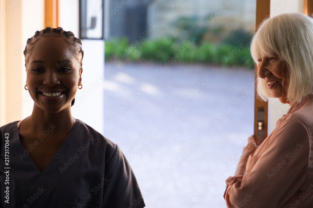 Naklejka premium African American caregiver standing in scrubs holding open front door while senior woman smiling