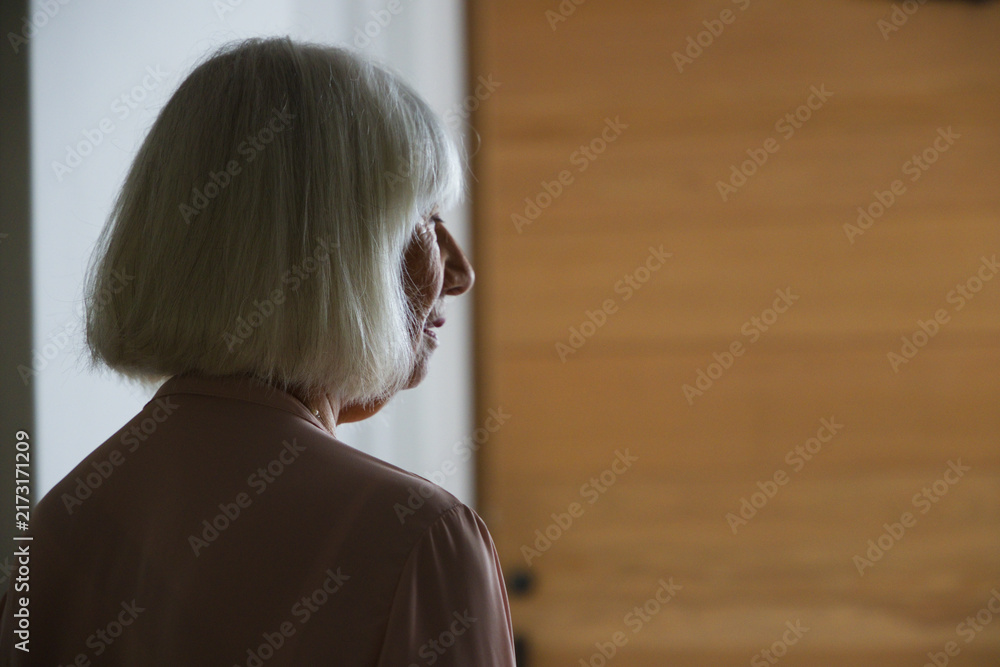Naklejka premium Senior female sitting in hallway by wooden door, knob and doorway, wearing pink blouse and necklace