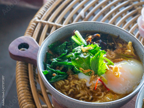 Fotografie Freshly cooked Asian noodle soup topped with soft poached egg, green vegetables, and crispy shallots served in a bowl on a rattan table