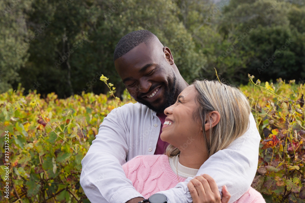 Naklejka premium Diverse couple embracing among vineyard rows during autumn, man wearing smartwatch and button-up