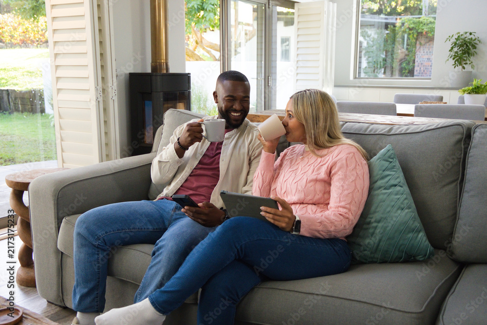 Naklejka premium Diverse couple sitting on gray sofa near sliding doors, holding mugs, using tablet and smartphone