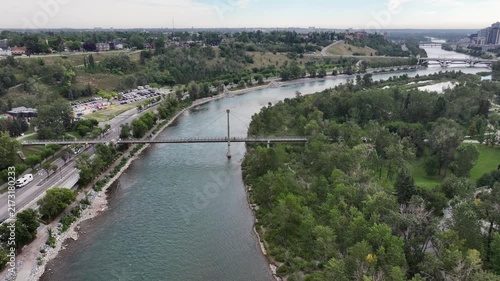 flying above Bow River next to Prince’s Island Park towards Centre St Bridge, while crossing Bow River Pathway Bridge with parts of Skyline of Downtown Calgary ( Alberta, Canada) on right side, video