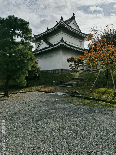 View of the traditional white turret of Nijo Castle with its dark tiled roof, stone foundation, and surrounding trees under a cloudy sky in Kyoto, Kyoto, Japan.