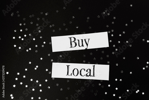 White paper strips with the message 'Buy Local' surrounded by star shaped confetti on a black background, promoting local shopping.