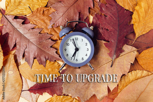 A blue alarm clock placed on a bed of autumn leaves with the text 'Time to Upgrade' in bold letters, suggesting change and renewal.