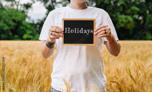 A person in a white t shirt holds a small chalkboard with the word 'Holidays' in a wheat field, suggesting a break or vacation theme.