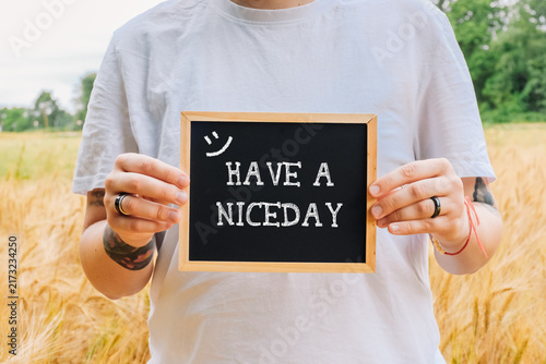 A person wearing a white t shirt holds a small chalkboard with the message 'Have a Nice Day' in a wheat field, suggesting positivity and well wishing.