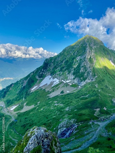 Fotografía Green alpine mountain rises above melting snow and cascading summer waterfall