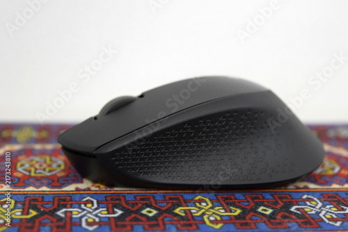 Foto Side profile view of a black ergonomic wireless optical mouse with textured rubber side grip resting on a colorful traditional carpet mousepad