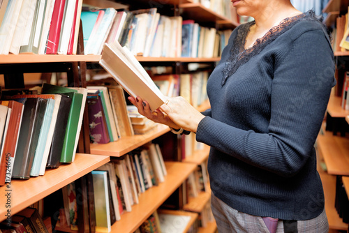A middle aged woman with glasses examines book in library. Bookshelves filled with various books surround her in school setting