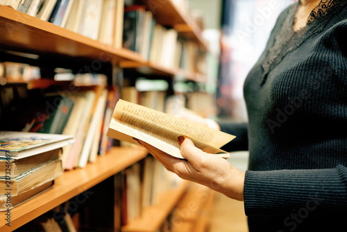 A middle aged woman with glasses examines book in library. Bookshelves filled with various books surround her in school setting