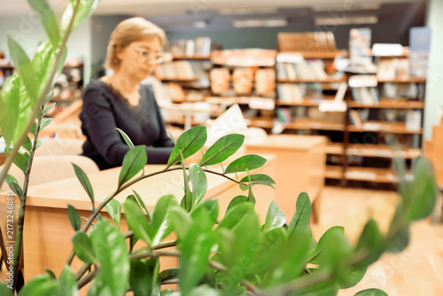 A middle aged woman with glasses reads book in the library. In school setting, she is surrounded by bookshelves filled with various books