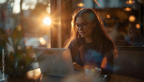 Young woman wearing glasses works on laptop in sunlit cafe during golden hour.