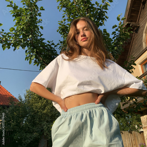 A young woman poses confidently in a bright outdoor setting, showcasing a casual summer look with natural surroundings.