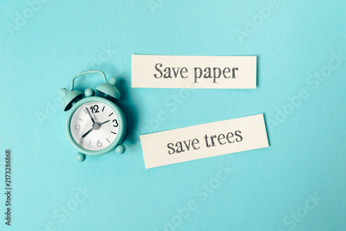 A vintage alarm clock next to paper strips with environmental messages 'Save paper' and 'save trees' on a blue background, emphasizing time and conservation.
