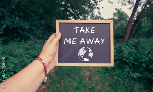 A hand holds a chalkboard with the message 'Take Me Away' and a globe illustration, set against a lush green outdoor background, suggesting a desire for travel and escape.