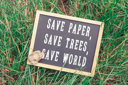 A chalkboard sign with the message 'Save Paper, Save Trees, Save World' placed on green grass, accompanied by a snail shell, emphasizing environmental conservation.