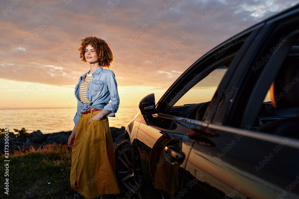 Naklejka premium Casual woman posing beside a car at sunset, wearing a denim shirt and yellow dress, capturing a relaxed lifestyle moment outdoors with warm light.