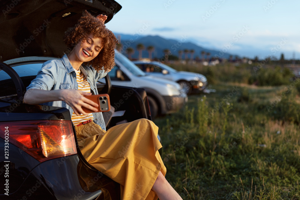 Fototapeta premium Smiling woman with curly hair sits on a car trunk taking a selfie. Outdoor parking lot beside a grassy field. Casual lifestyle, travel, wellness vibe and carefree mood.