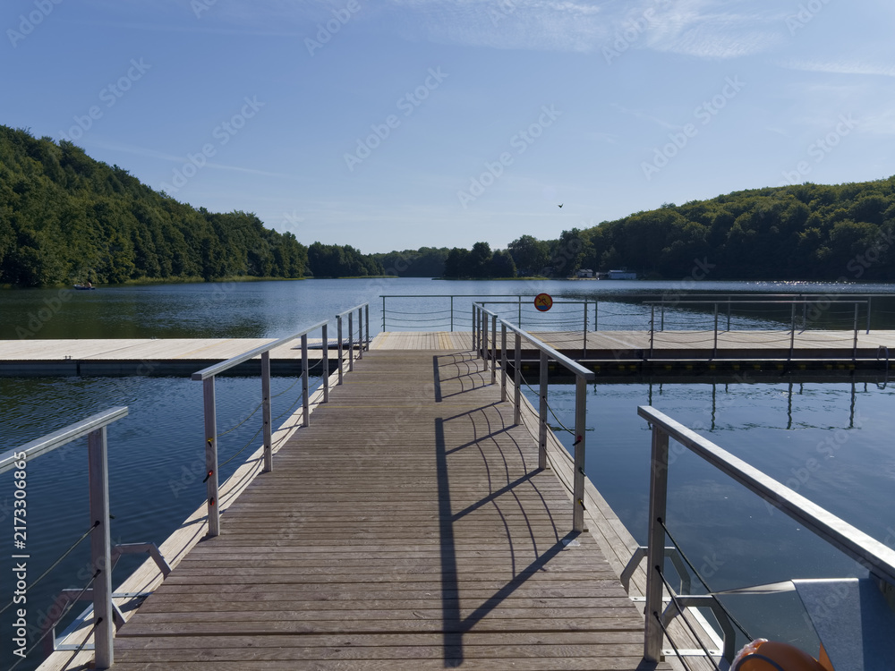 Fototapeta premium Visitors walk on a wooden pier by a river surrounded by trees on a sunny day in the countryside