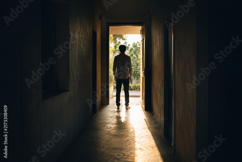 Man silhouette walking towards bright sunlight doorway at end of dark hallway