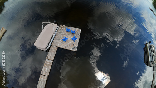 Obraz na plátně A top down close up wide angle drone shot highlights a wooden lake dock featuring blue adirondack chairs and a covered pontoon boat on dark reflective water on a sunny day