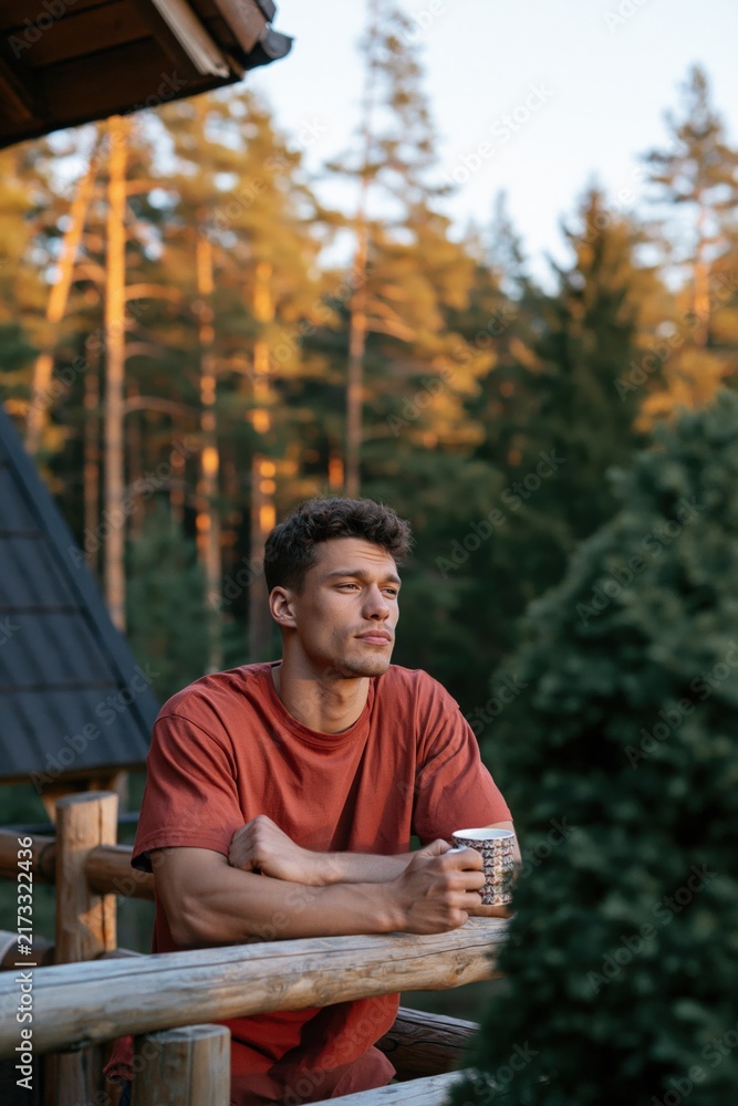 Fototapeta premium Young man on log cabin balcony holding a mug at sunset among pine trees for outdoor lifestyle background and travel banner