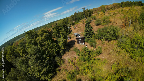 Obraz na płótnie A high angle wide shot looks down over a small dark tiny house cabin nestled in a grassy clearing on a rugged woodland hill under a bright blue sky filled with clouds