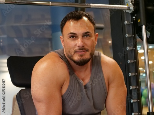 Cuadro en lienzo Close-up portrait of a sweaty muscular man smiling gently while looking at the camera, resting between workout sets in a fitness gym
