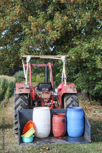 A red tractor transports blue, white, and red water barrels in a rear bucket to irrigate crops during a severe heatwave and drought in the hot summer of 2026. Realistic portrait photograph.