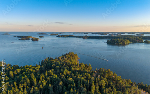 Aerial view of a boat leaving a wake through the Helsinki archipelago at golden hour, with forested islands and calm blue water, Finland