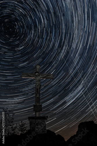 Star trails circling the night sky above the summit crucifix on Kleiner Sonnstein near Traunkirchen, Upper Austria, with Alpine silhouettes and aircraft light trails.