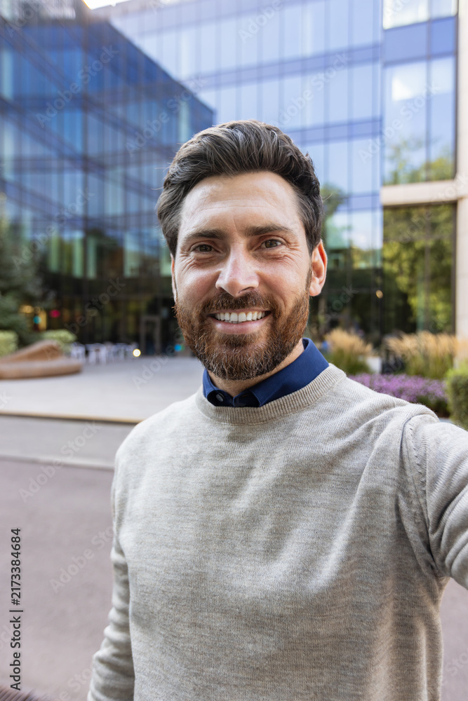 Fototapeta premium Vertical close-up photo of a young man standing outside a building, holding a camera in his hand and chatting online
