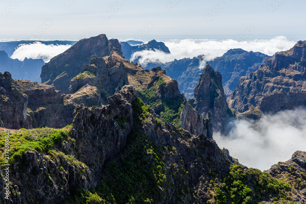 Fototapeta premium Volcanic peaks dominate Madeira's rugged landscape. Clouds drift between cliffs at Pico do Areeiro. Green vegetation contrasts dark rocky terrain under blue sky.