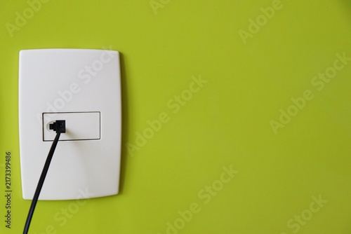 Black telephone line cable plugged into a white phone wall socket plate on a green wall. Landline telephone connection and telecommunications.