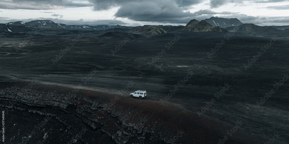 Naklejka premium Aerial view of a lone 4x4 vehicle crossing the remote volcanic highlands of Iceland, surrounded by dark lava fields, rugged mountains, snow patches and dramatic storm clouds.