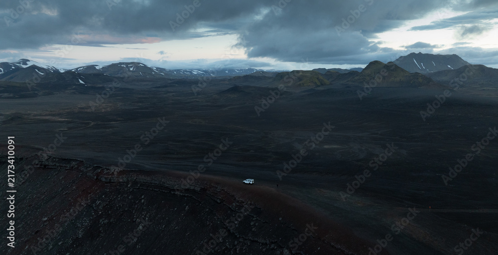 Naklejka premium Aerial view of a lone 4x4 vehicle crossing the remote volcanic highlands of Iceland, surrounded by dark lava fields, rugged mountains, snow patches and dramatic storm clouds.