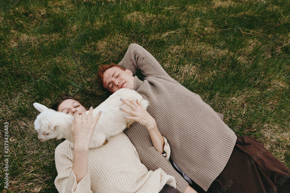 Naklejka premium Casual outdoor moment with two friends and a playful goat kid lying on grass, cozy sweaters, soft light, relaxed smiles, natural colors, rustic atmosphere, lifestyle scene outdoors
