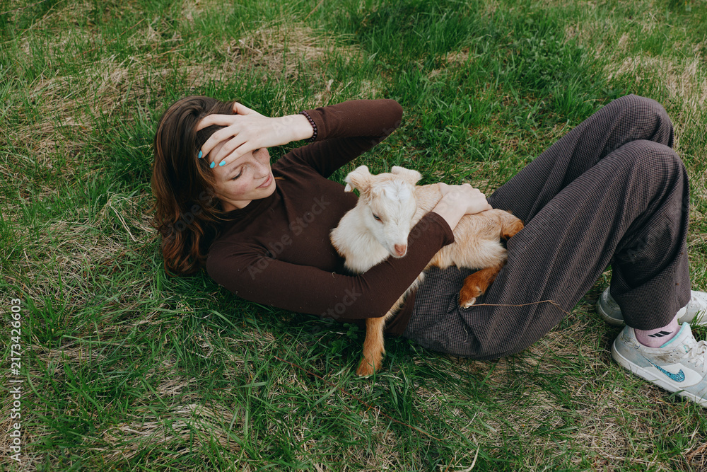 Naklejka premium Relaxed woman lying on grass outdoors with a playful goat kid resting on her chest, sunlit moment, casual clothes, gentle breeze, natural colors, tranquil countryside vibe