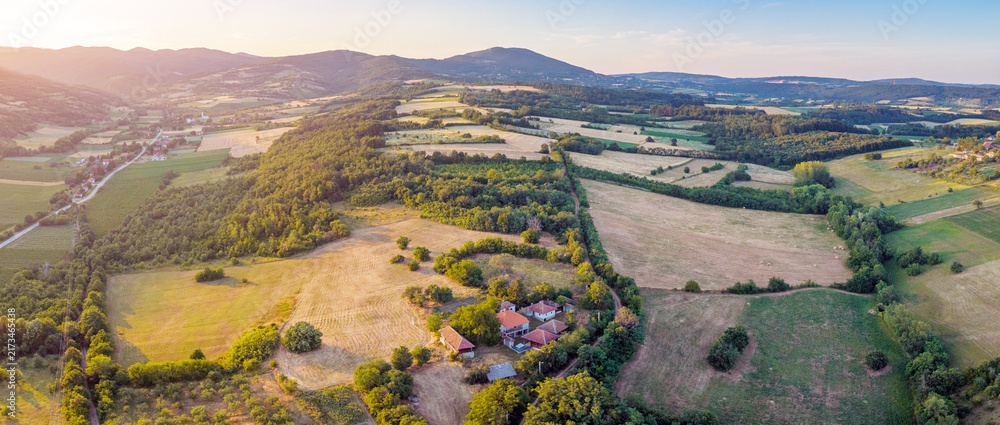 Naklejka premium Aerial panoramic view of rolling countryside farmland with red-roofed homestead at golden sunset. Rural escape concept.