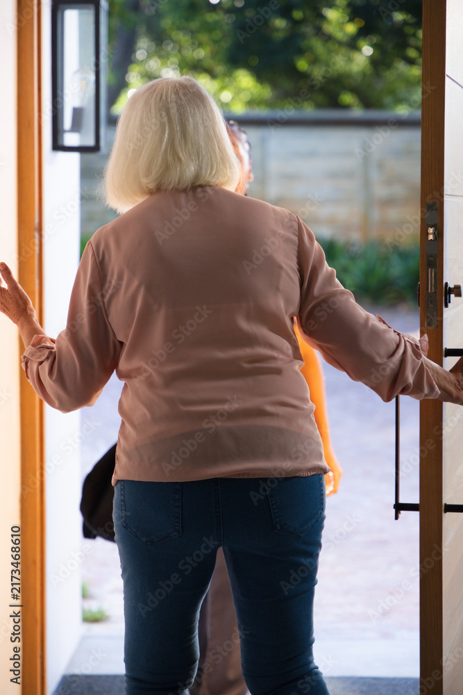 Naklejka premium Senior woman in pink blouse holding door in doorway with metal latch greeting woman carrying bag