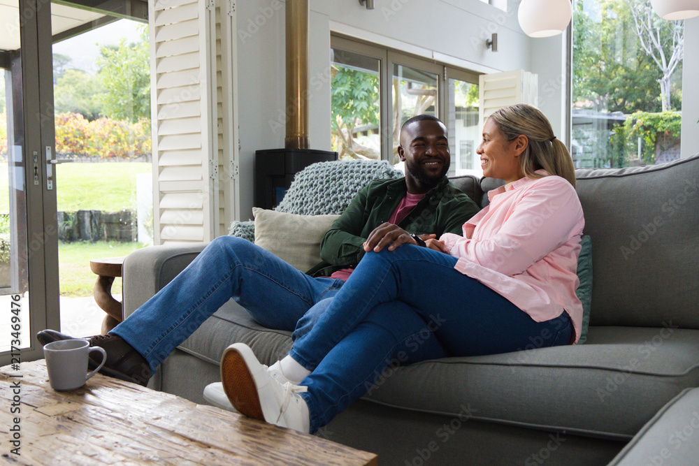 Naklejka premium Diverse couple sitting on grey sofa in living room holding hands beside coffee table with mug