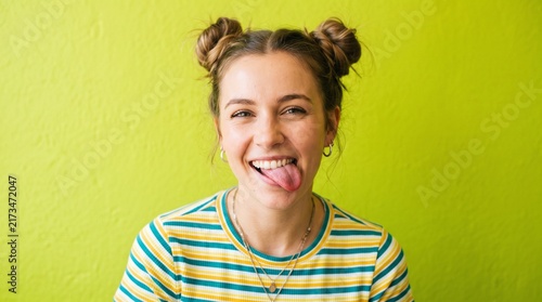 Playful happy young woman with space buns hairstyle sticking out tongue against bright vibrant lime green background