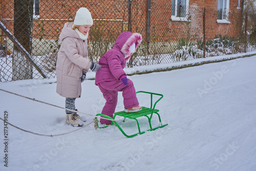 Older girl helping toddler sister to step on green sled outdoors in winter, Two small children playing with sleds on a snowy street