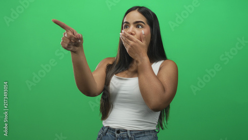 Fotografie Hispanic woman pointing finger with left hand and covering mouth with right hand in green studio; surprise discovery