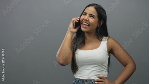 Картината върху платно Young hispanic woman holds phone to ear smiling in a grey studio wearing a white tank top and jeans; happiness connection