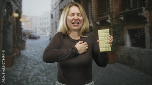 Fotografía Blonde woman clutching chest holds large white chocolate bar in right hand on a cobblestone street with stone buildings; heartattack pain