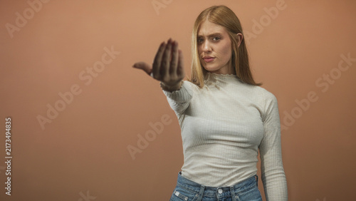 Cuadro en lienzo Young redhead woman in white long sleeve top and jeans beckons with outstretched hand toward camera in studio; assertive invitation