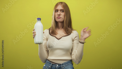 Obraz na plátně Young woman holds milk bottle and waves her hand with a displeased expression in a studio with yellow backdrop; disgust rejection